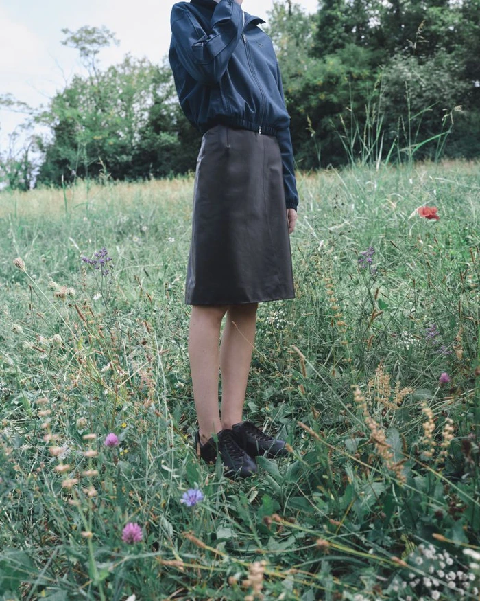 Model standing outdoors in a wild grassy field wearing a blue zip jacket, dark midi skirt, and black sneakers.