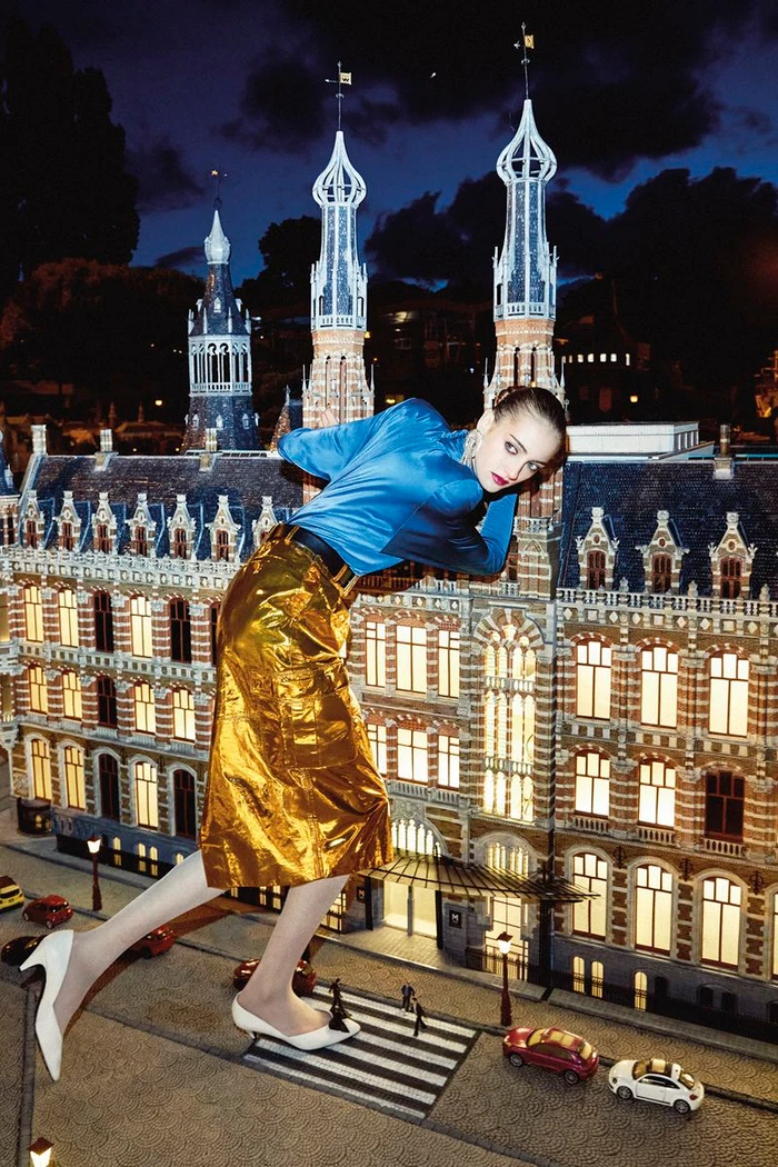 Model Frauke Nijs in blue top and metallic gold skirt posed against a miniature cityscape