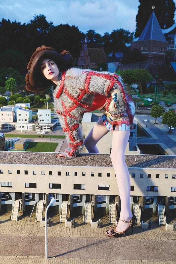Model Frauke Nijs wearing a textured furry jacket and wide hat, posed over a miniature village model for D Repubblica