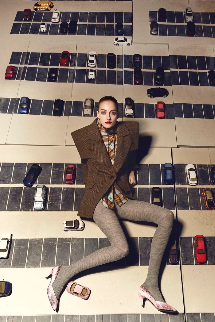 Model Frauke Nijs in a structured brown blazer and grey tights posed against a miniature parking lot background for D Repubblica