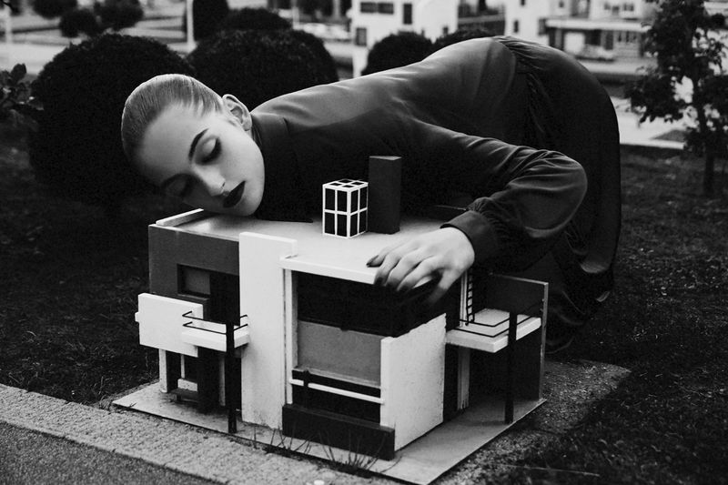 Model Frauke Nijs leaning over a miniature modernist architectural model in a surreal editorial photograph.