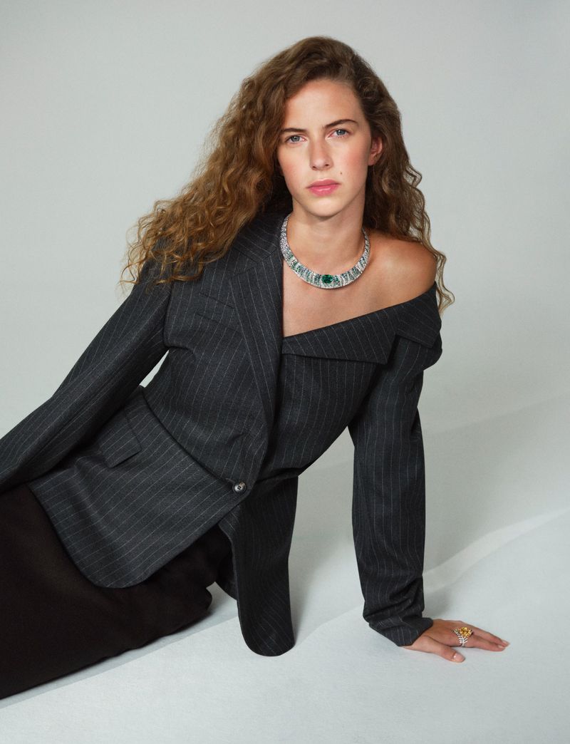 Model with long curly hair wearing an off-the-shoulder pinstripe blazer and statement necklace against a light grey seamless background
