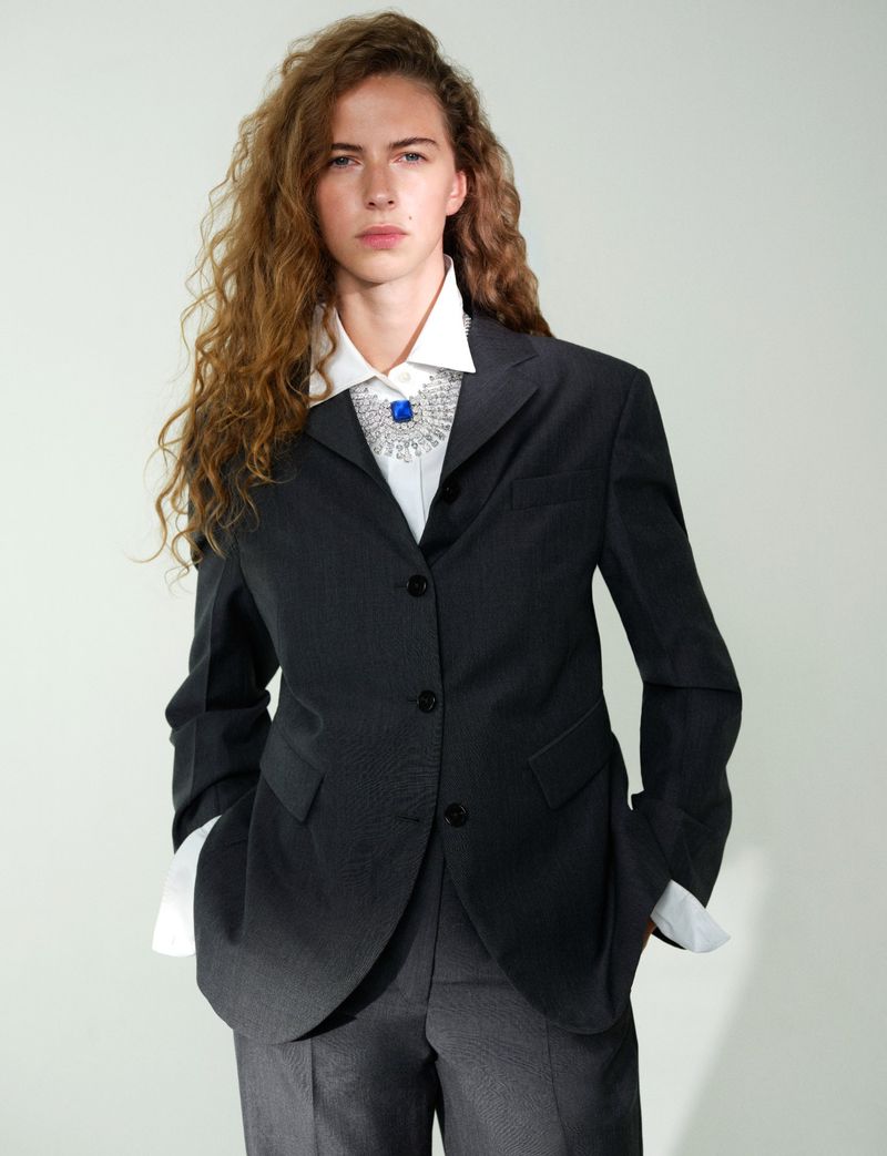 Model with long curly hair wearing a dark grey blazer, white shirt, and jeweled necklace against a grey backdrop