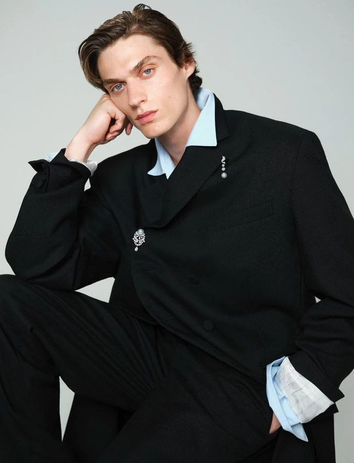 Male model in a black suit jacket and light blue shirt resting head on hand against a neutral grey background.