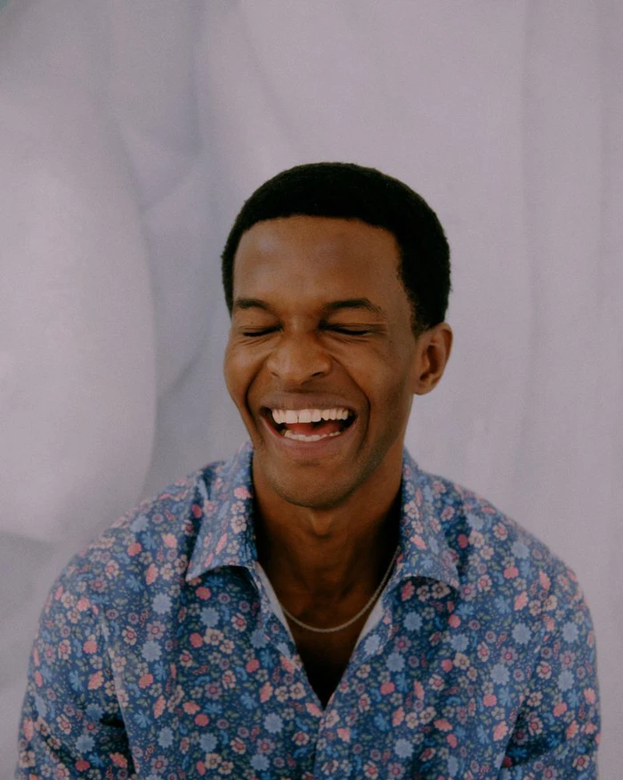 Close-up portrait of a laughing Black man wearing a blue floral shirt against a pale backdrop