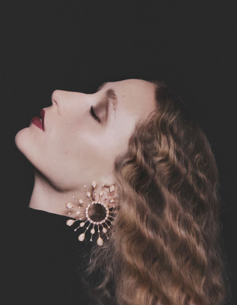 Profile portrait of a woman with wavy hair, dark lipstick, and ornate sunburst earrings against a dark background in a studio setting.