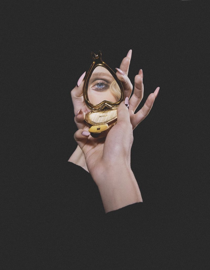 Hand holding a heart-shaped mirror with a blue eye reflection against a dark backdrop, manicure and gold accents visible.