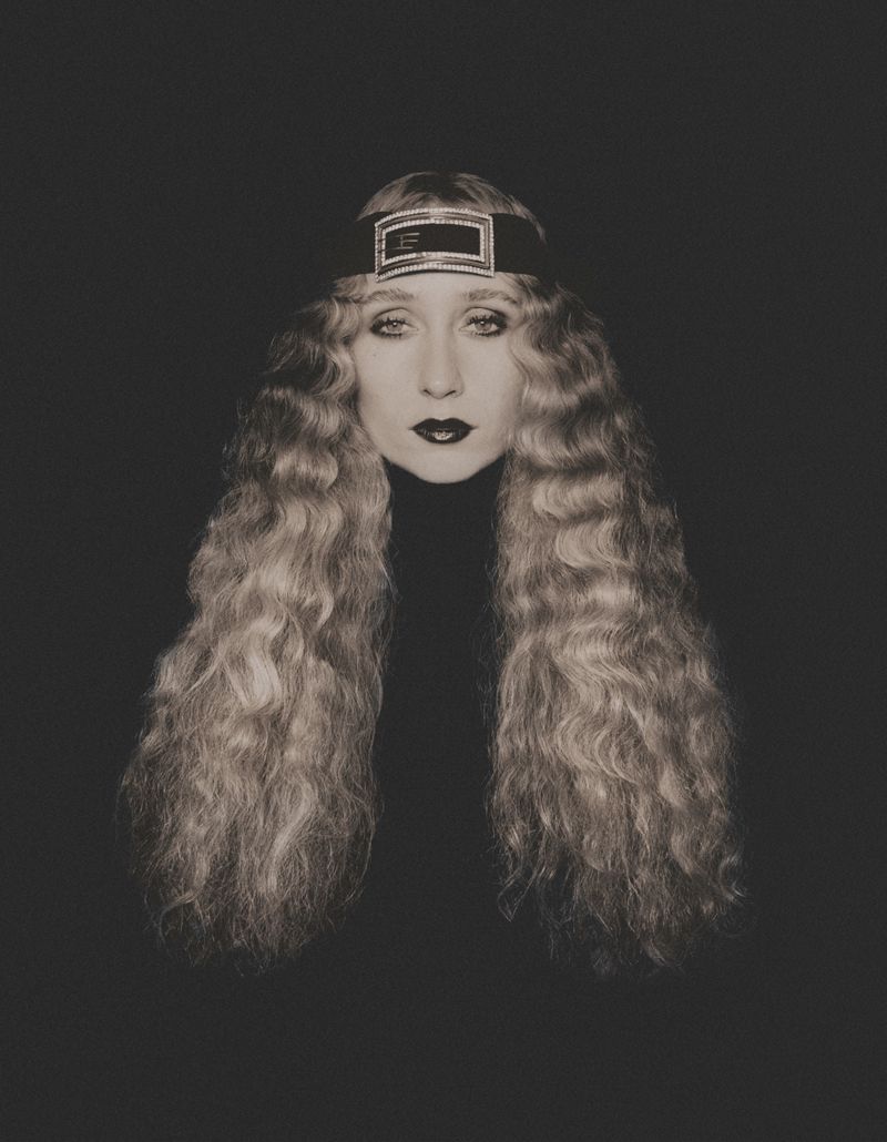 Portrait of a woman with long, curly blonde hair and a black headband, wearing a black turtleneck against a dark background.