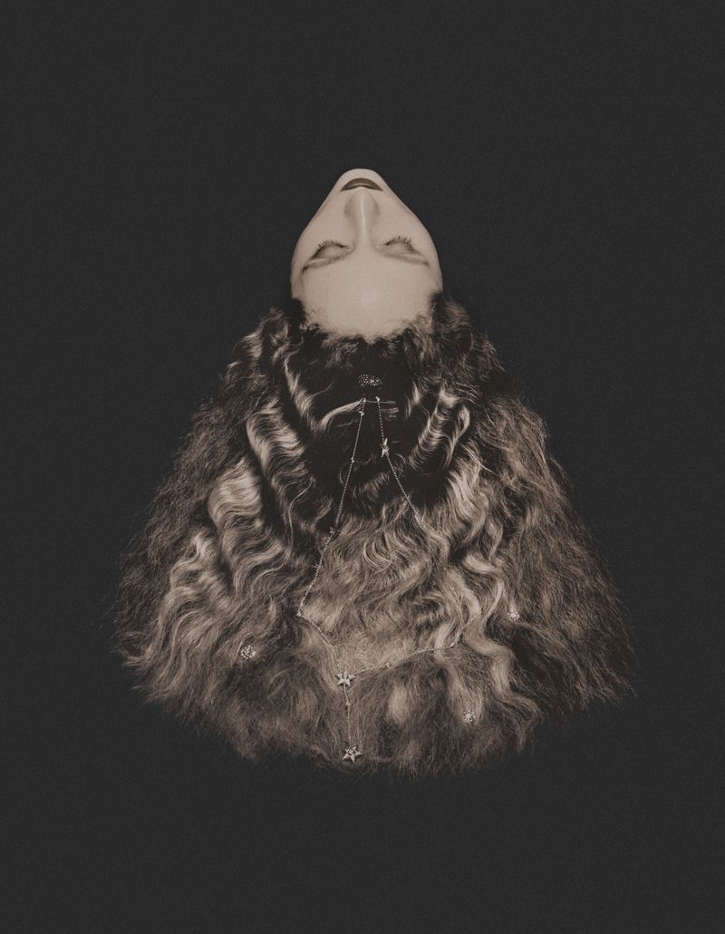 Monochromatic studio portrait of an adult female with curly brown hair, pale skin, and a necklace with star pendants, posing against a dark backdrop.