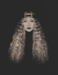 Portrait of a woman with long crimped blonde hair wearing a dark turtleneck and headband against a dark background