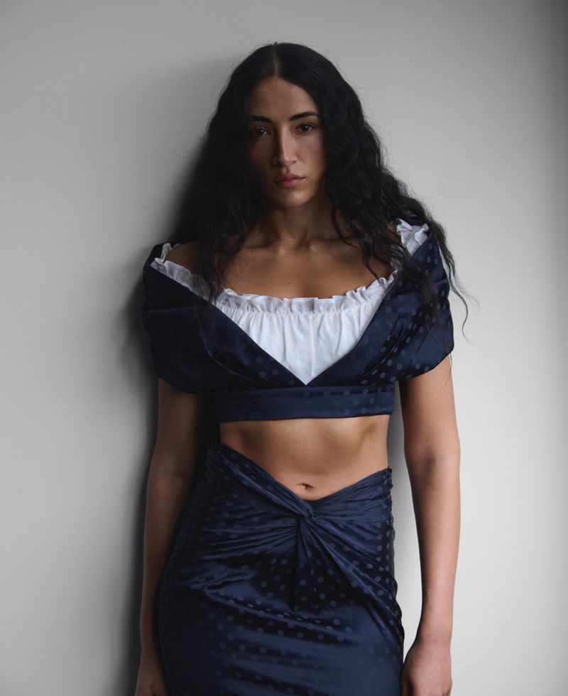 Portrait of a woman in a two-piece blue patterned outfit with a white ruffled top, posing in front of a textured grey wall.