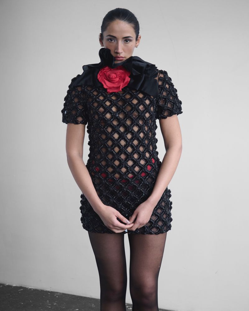 Woman in a black textured mini dress with a large red flower, posing against a plain wall, showcased in a studio-style fashion shoot.