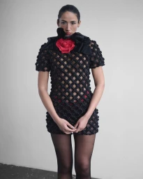 Model Cora Corré wearing a black netted dress with a red rose collar against a plain studio background.