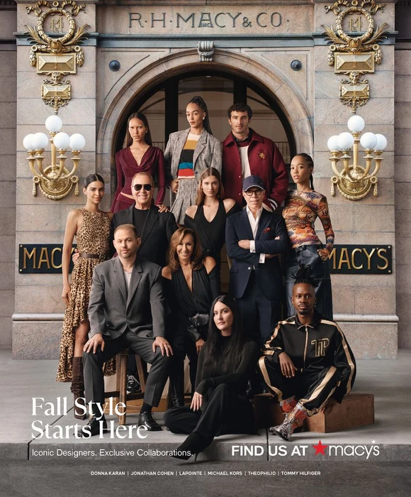 Group portrait of fashion designers and models posing in front of a historic Macy's storefront with stone arches and gold signage.
