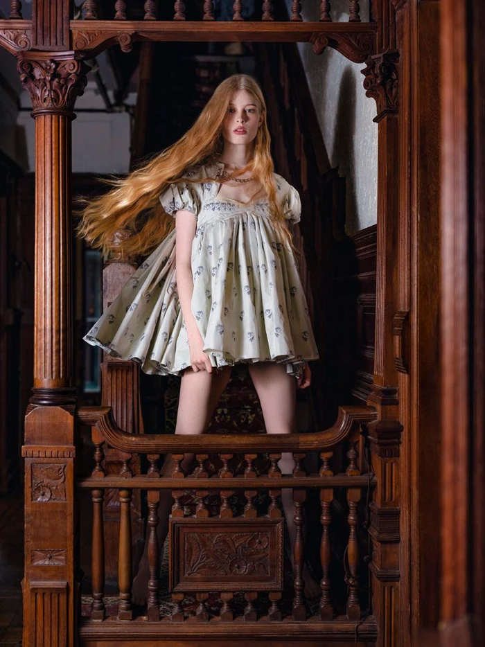 Red-haired model in a patterned babydoll dress standing behind ornately carved wooden staircase balusters