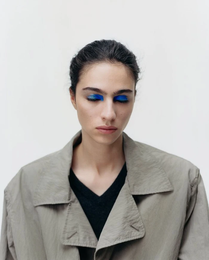 Close-up portrait of model Loli Bahia wearing blue eye makeup, a black top, and a grey coat against a plain white background.