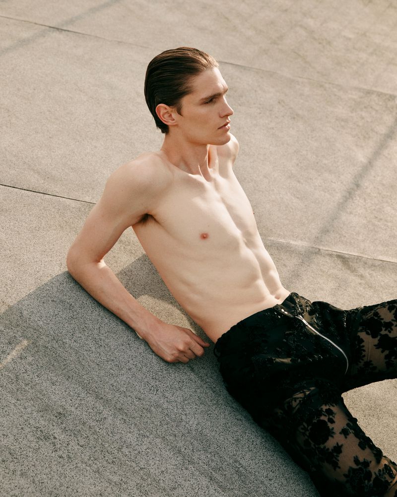 Shirtless male fashion model in patterned black pants laying on concrete outdoors in portrait layout with copy space for visual marketing.