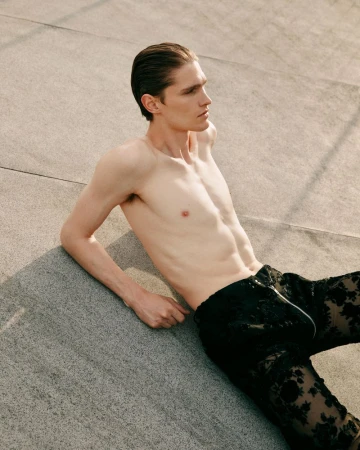 Shirtless male fashion model in patterned black pants laying on concrete outdoors in portrait layout with copy space for visual marketing.