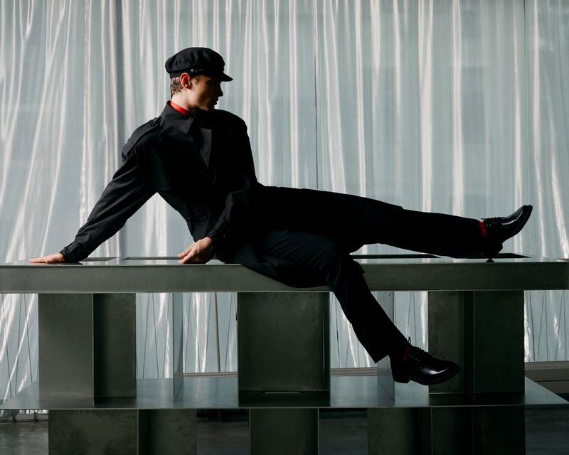 Young man in a black outfit and cap poses on a metallic surface against a translucent white background in an indoor setting.