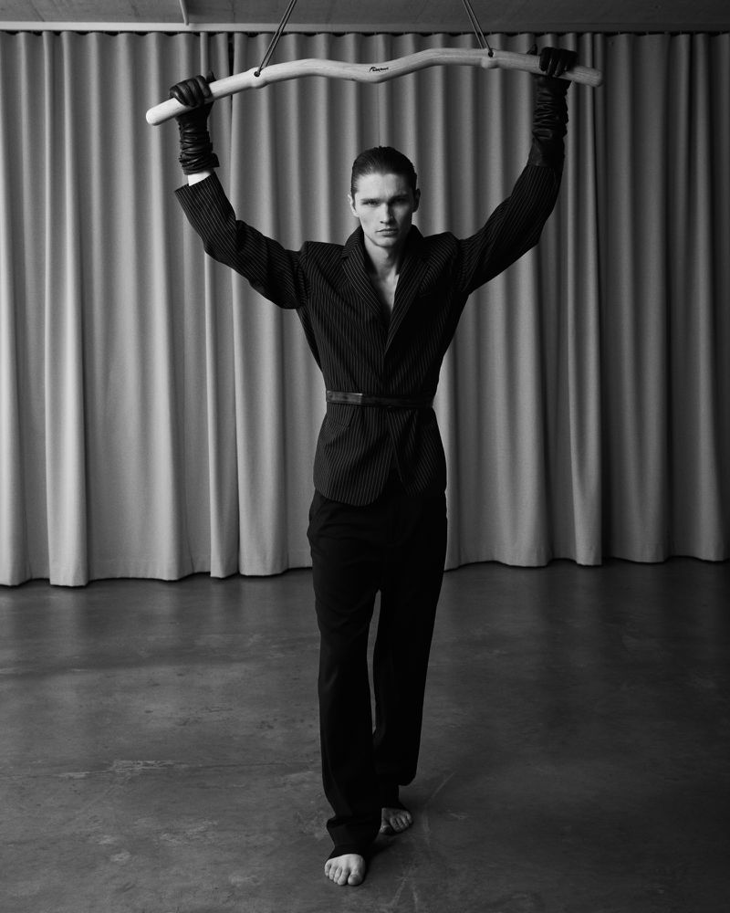 Striking black-and-white fashion portrait of a slender man with bare feet holding up a branch in front of a neutral curtain backdrop.