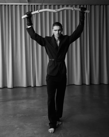 Striking black-and-white fashion portrait of a slender man with bare feet holding up a branch in front of a neutral curtain backdrop.