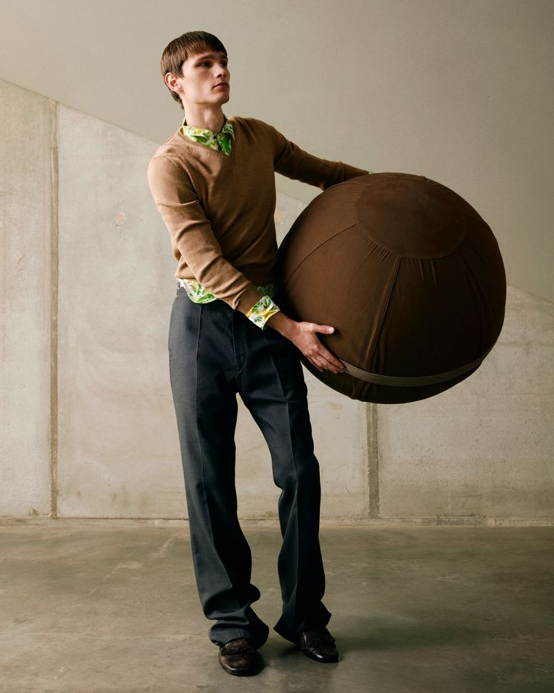 A slender male model holds a large brown spherical object in an indoor photo studio, wearing a brown sweater and wide-leg grey trousers.