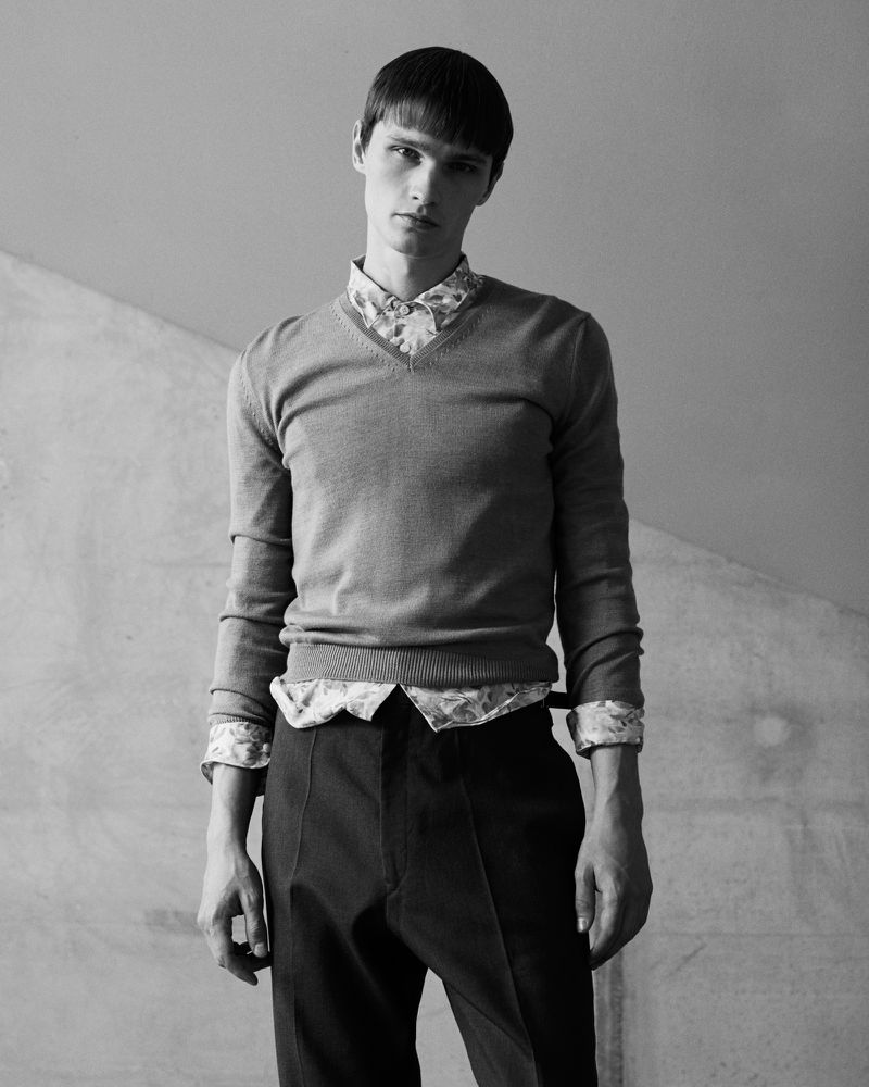 Portrait of a young adult male wearing a grey sweater and patterned shirt against a grey wall, looking directly at the camera.