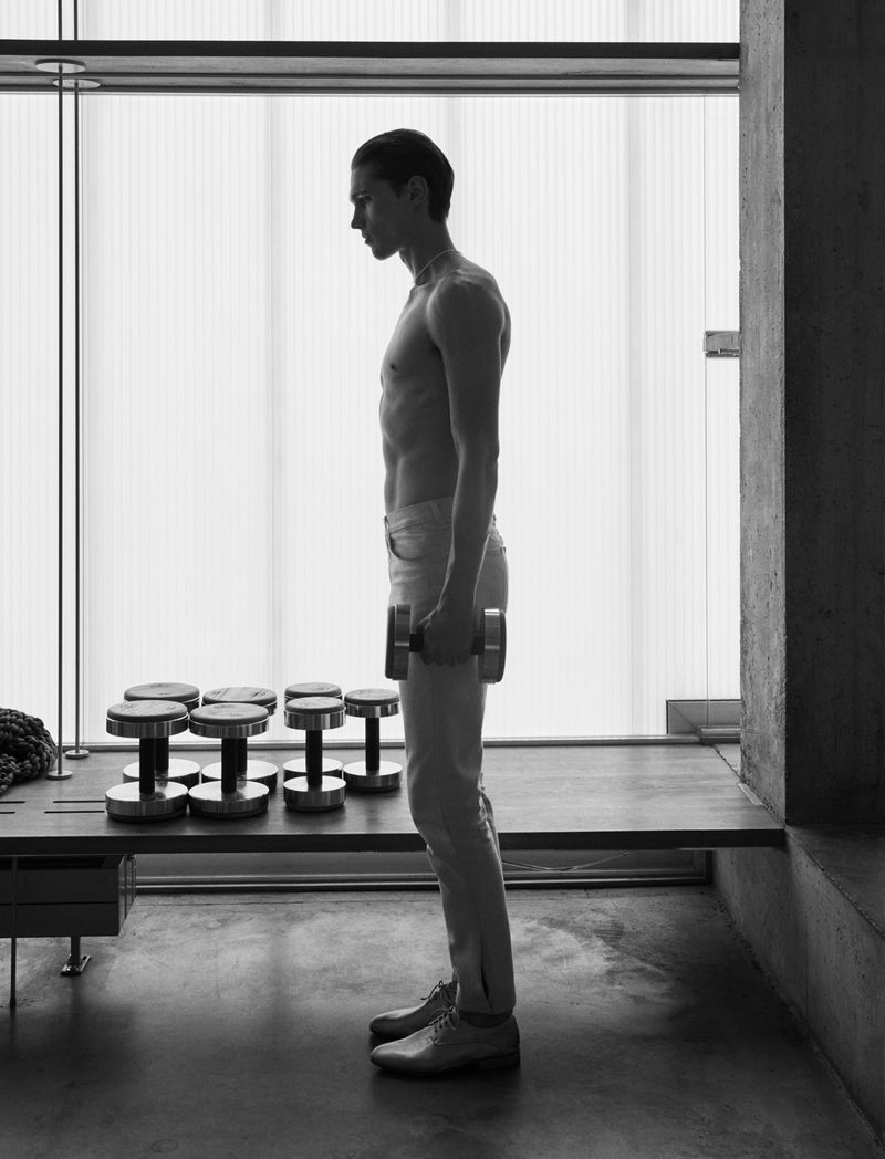 Monochrome studio shot of a shirtless adult male lifting a dumbbell, with additional weights on a modern table against a window backdrop.