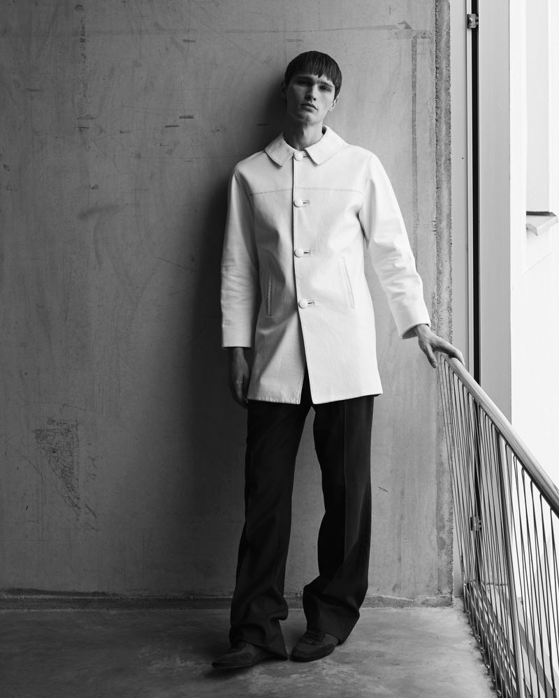 Monochrome portrait of a young adult male fashion model wearing a white coat and dark pants, leaning against a railing near a concrete wall.