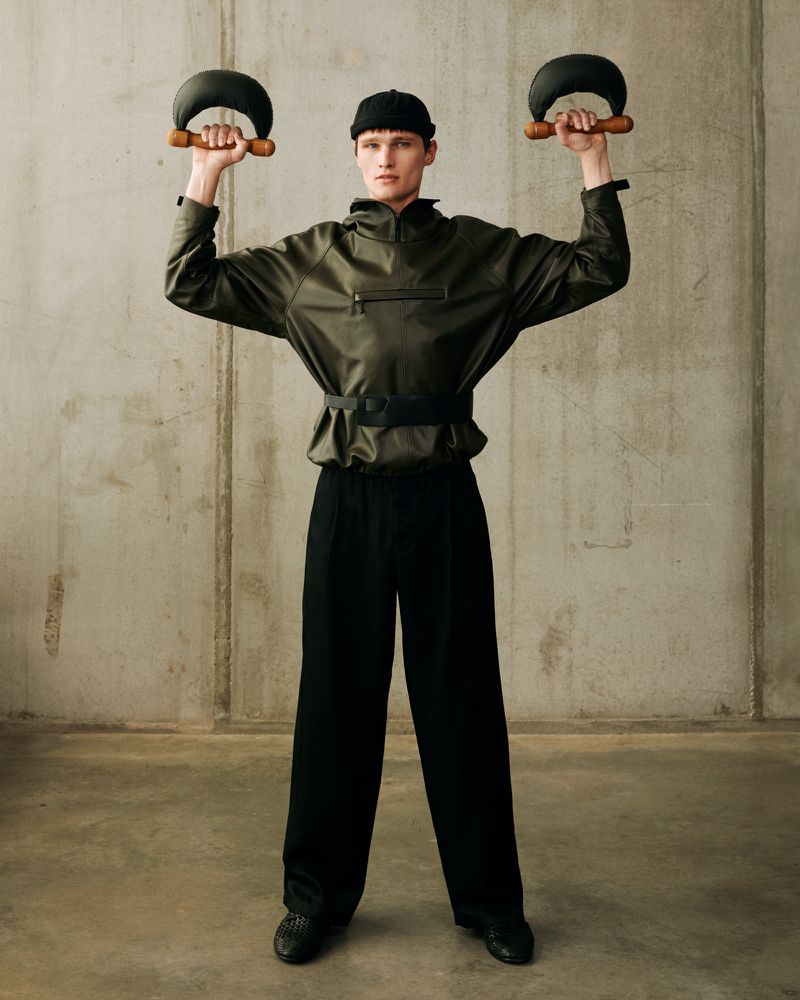 Man in green jacket and black hat lifting kettlebells in photo studio. Fashion model poses with weights for fitness editorial shoot.