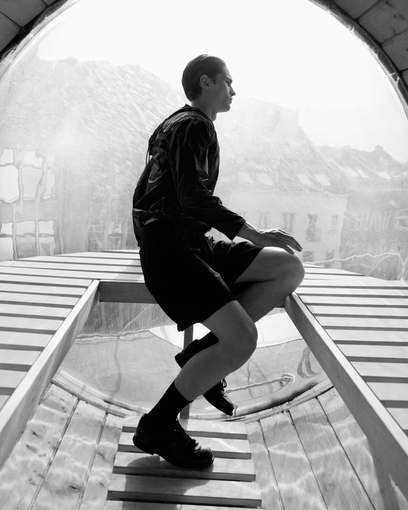 Black and white photo of a man in shorts and jacket climbing steps inside a curved, transparent structure with mountains visible outside.