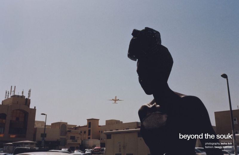 Silhouette of an adult wearing a headwrap in front of city buildings with an airplane visible in the sky on a sunny day.