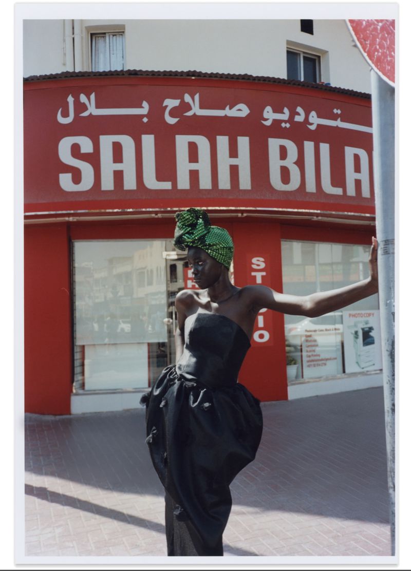 Woman in black dress and green headscarf poses in front of red building with 'SALAH BILAL STUDIO' sign in urban outdoor setting.