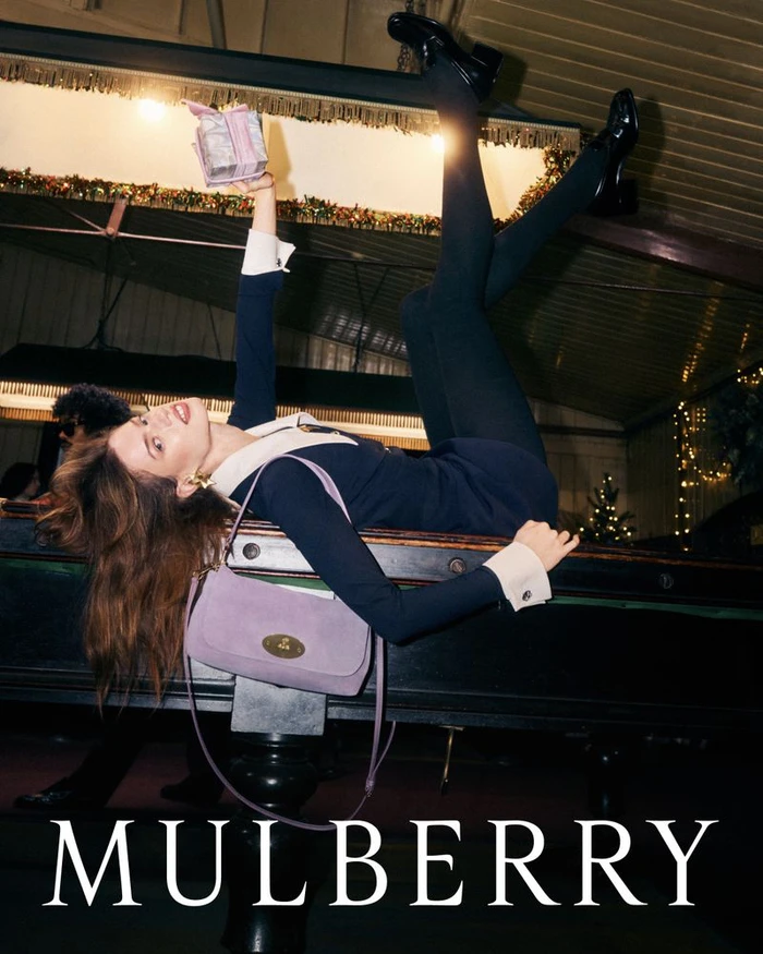 Model Lily McMenamy lying on a pool table holding a gift box, wearing a dark dress with white cuffs and a purple shoulder bag for Mulberry.