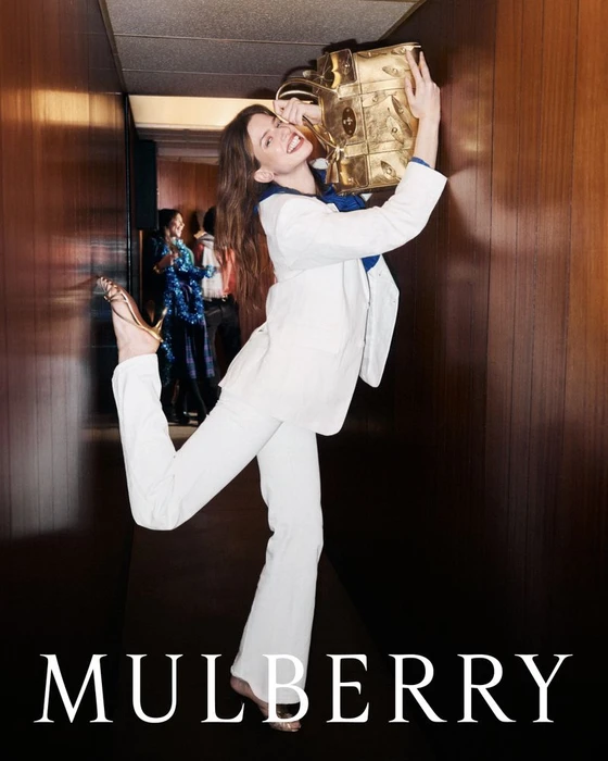 Model Lily McMenamy in white suit holding a gold bag in a wood-paneled hallway for Mulberry Festive 2026 campaign.