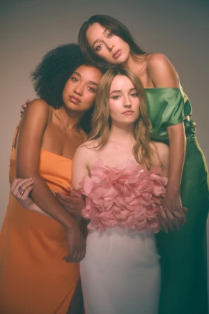 Three women in colorful gowns posing in a photo studio. One wears peach, one orange, and one green against a neutral backdrop.