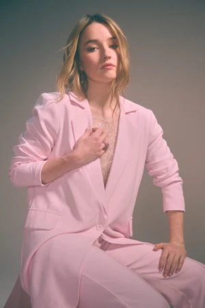Kaitlyn Dever wearing a pink blazer and trousers seated against a neutral studio background for Apple Cider Vinegar publicity portraits.