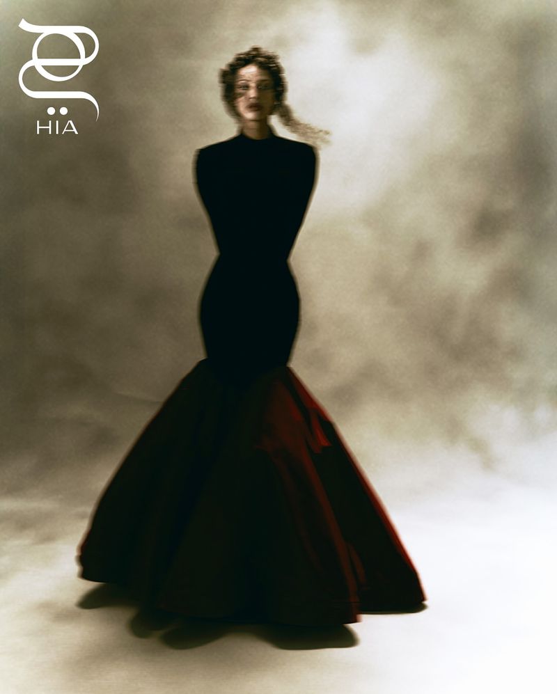 Fashion editorial shot of a woman in a black and red gown with hands behind her back, posing in front of a mottled grey background.