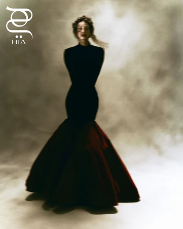 Fashion editorial shot of a woman in a black and red gown with hands behind her back, posing in front of a mottled grey background.