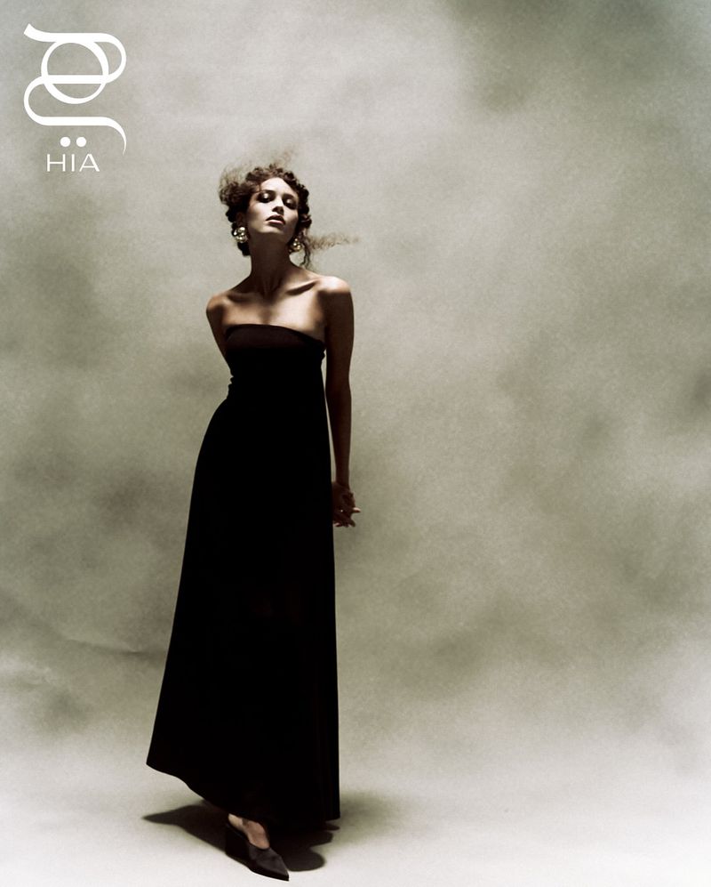 Elegant woman in strapless black dress posing in a photo studio with soft lighting against a textured grey backdrop, fashion editorial.