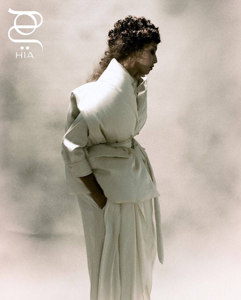 Side profile of adult woman in long white belted coat with hand in pocket against blurred background in studio, fashion editorial.