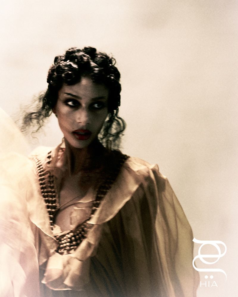 Elegant portrait of an adult woman in a sheer, ruffled dress with vintage-inspired hair, a beaded collar, and red lipstick against a light background.