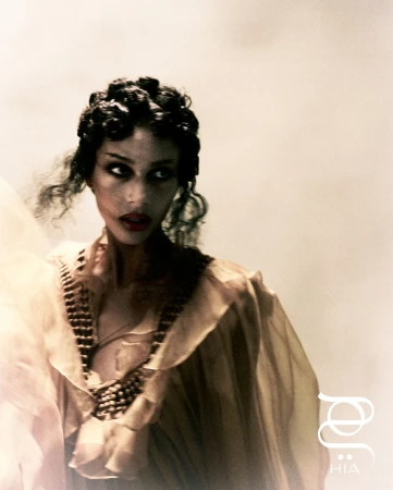 Elegant portrait of an adult woman in a sheer, ruffled dress with vintage-inspired hair, a beaded collar, and red lipstick against a light background.