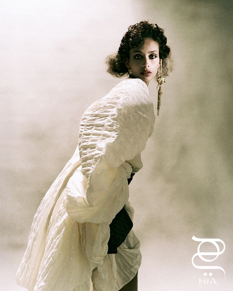Woman poses in a large, ruffled white gown with statement earrings against a soft grey backdrop in a fashion photography setting.