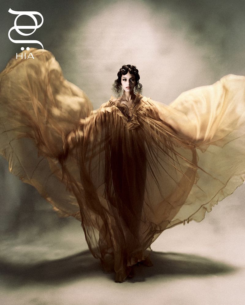 Woman in flowing gold gown poses with arms outstretched in a fashion portrait with soft lighting against a neutral gradient background.
