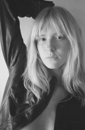 Black and white portrait of a young woman with freckles and long blonde hair, wearing a leather jacket in front of a plain background.