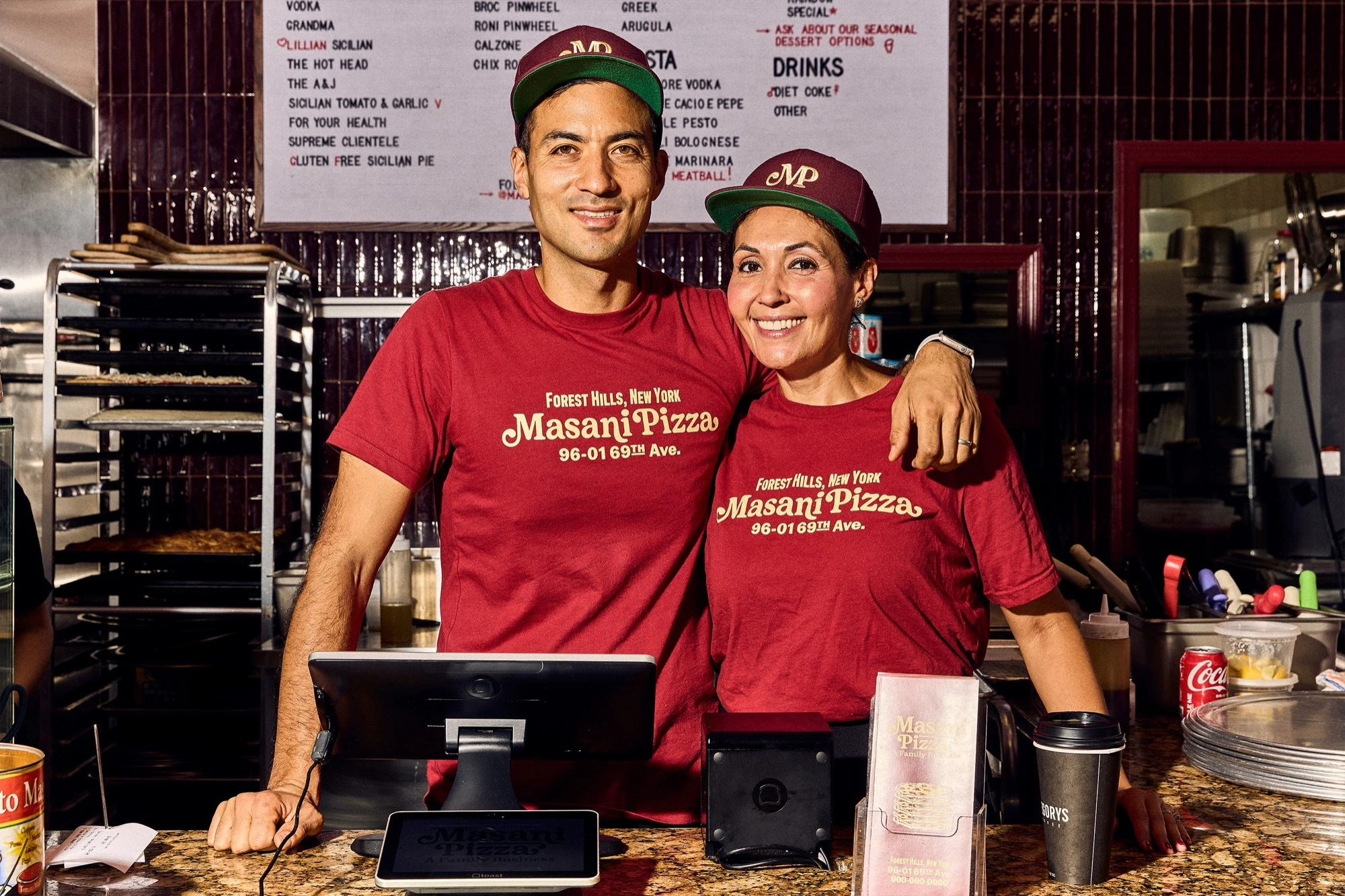 Two people wearing branded maroon t-shirts and baseball caps posing behind a restaurant counter for Masani Pizza.