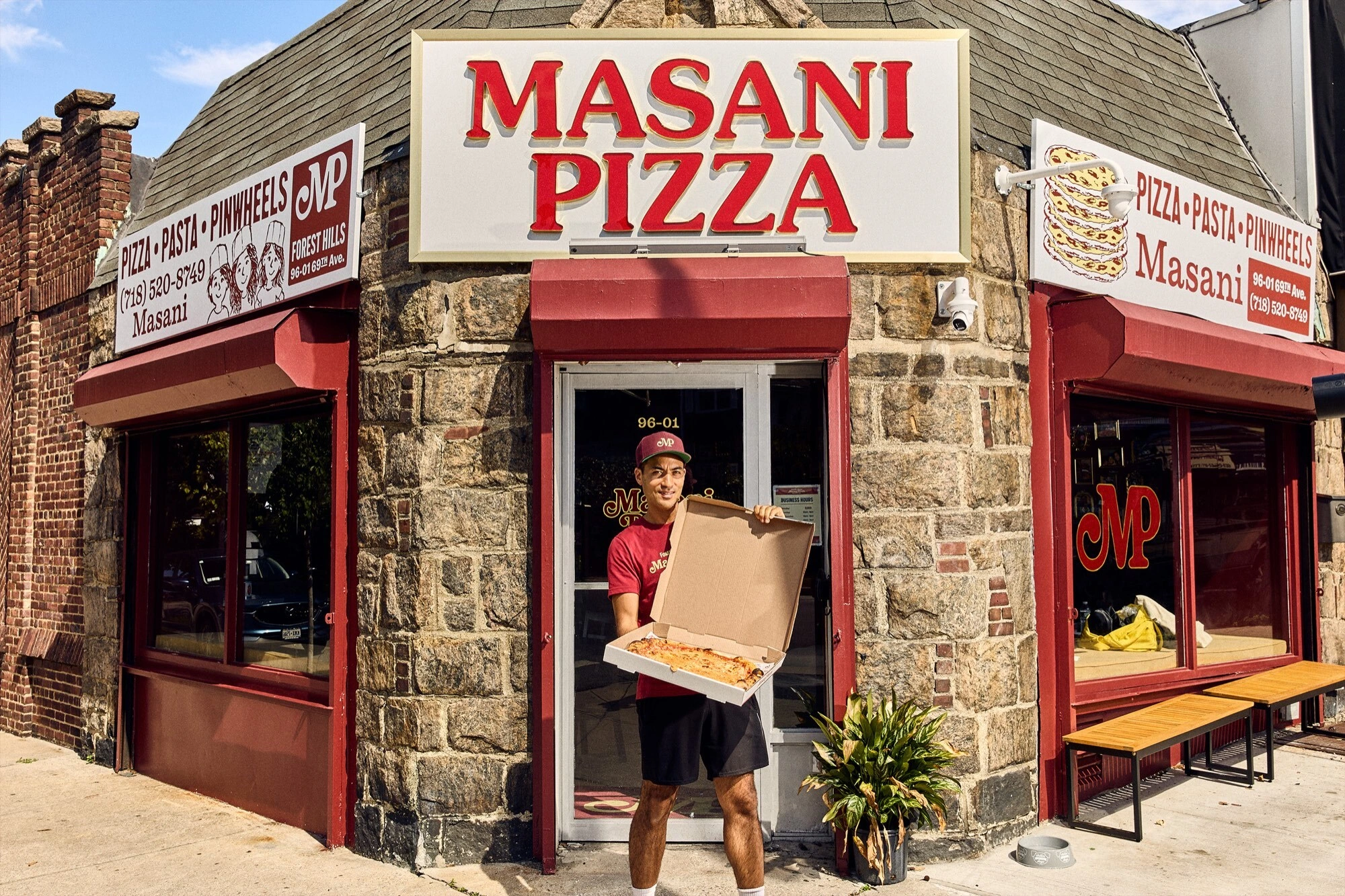 Man holding open pizza box standing in front of Masani Pizza storefront with stone facade and red awnings