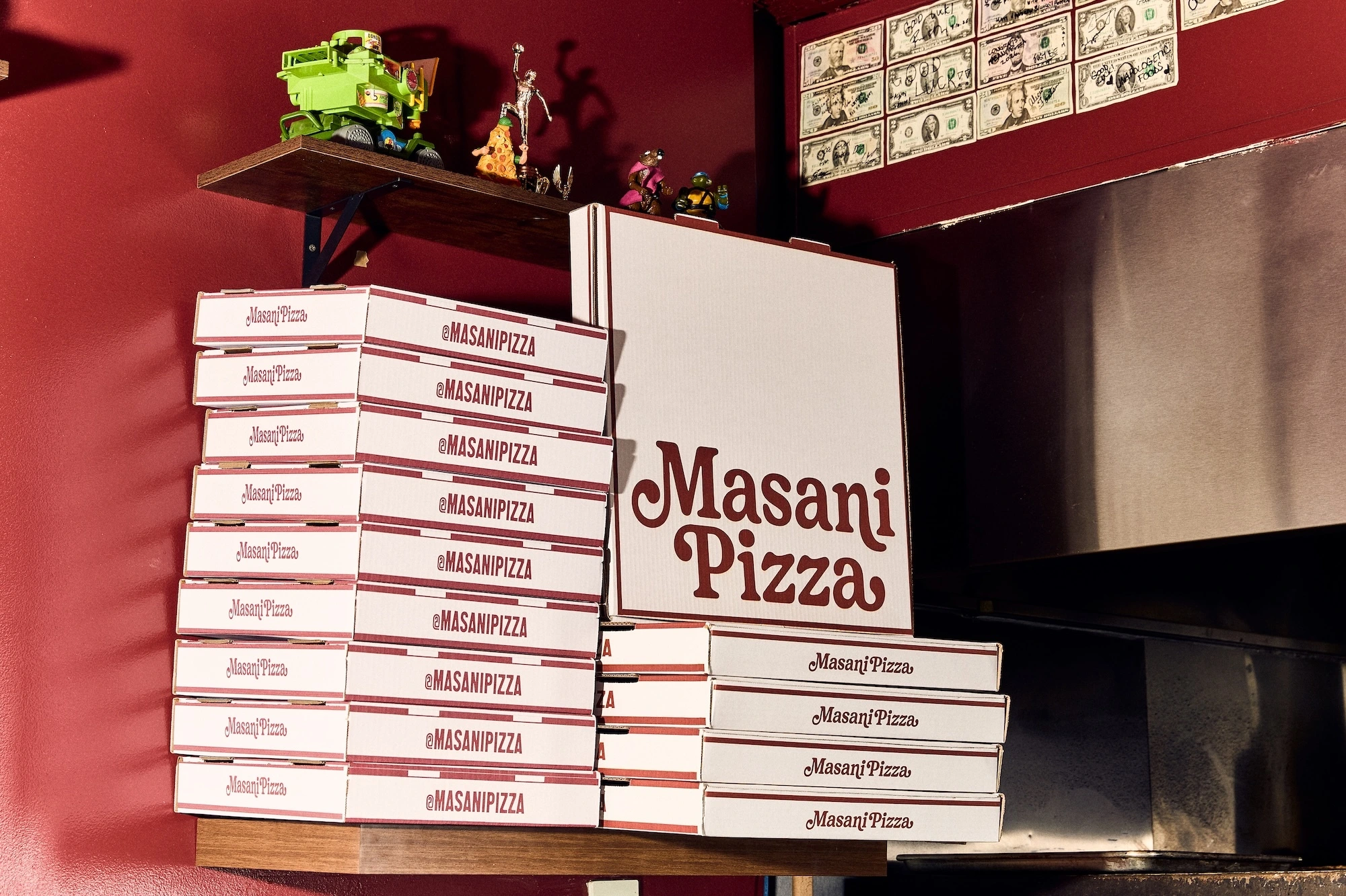 Stacks of branded Masani Pizza boxes displayed on wooden shelves against a deep red wall with framed dollar bills.