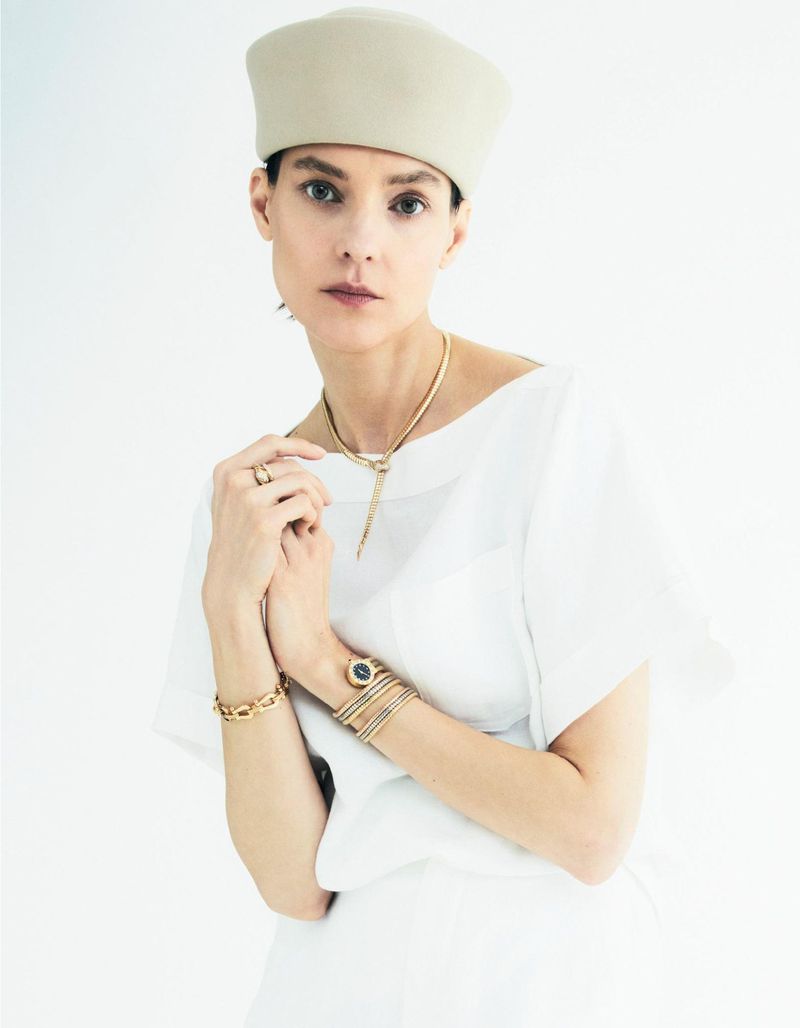 Elegant woman wearing jewelry and a hat poses against a white background. She wears a white blouse, gold jewelry and multiple bracelets.